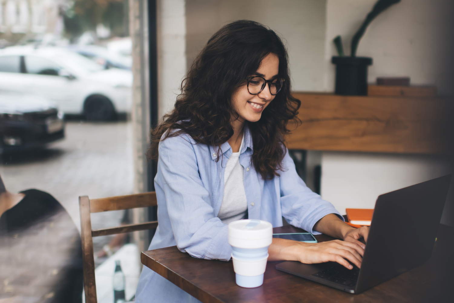 A young woman with long, curly dark hair and glasses sits at a cafe table, smiling while typing on her laptop