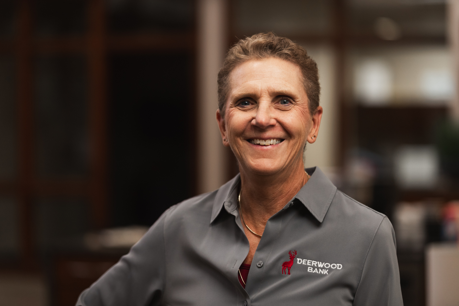 Headshot of a smiling woman with short, light brown hair wearing a grey button-up shirt featuring the Deerwood Bank logo