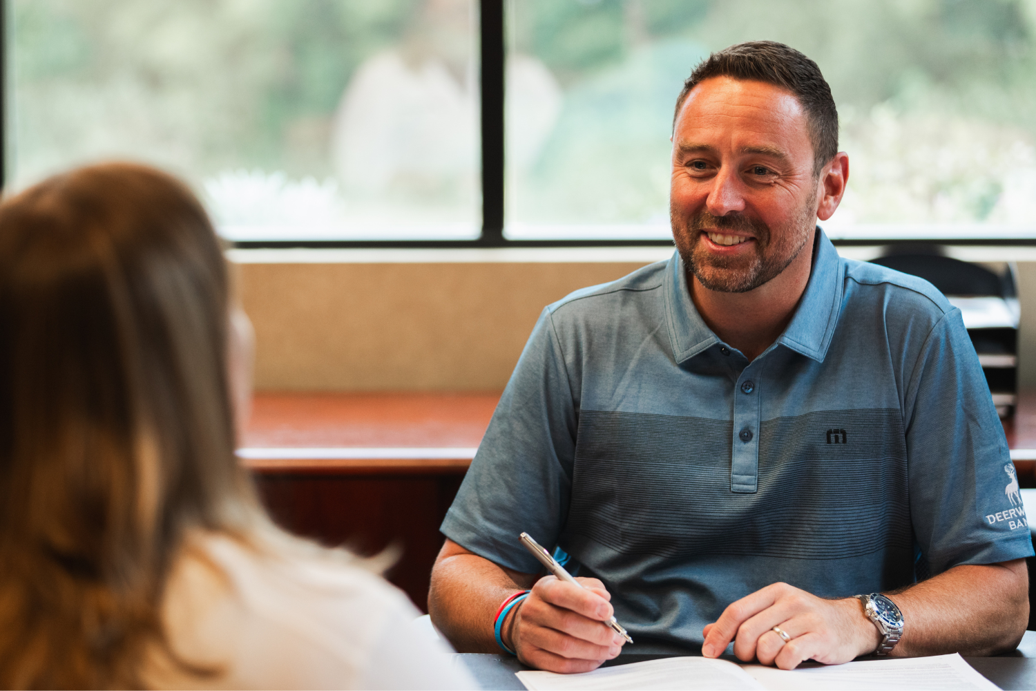 A smiling man with short hair and a beard sits at a desk, holding a pen and looking at a woman across from him during a meeting or consultation