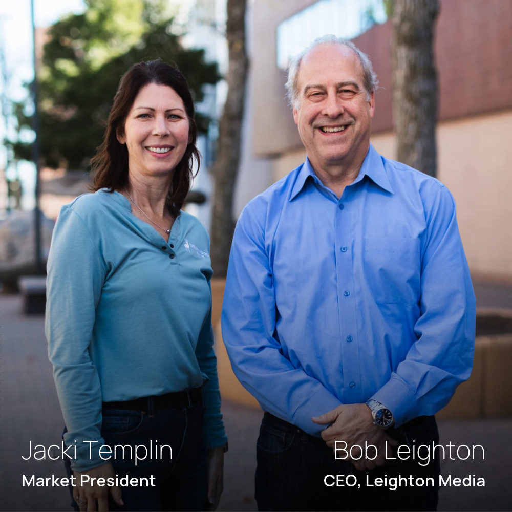 A smiling woman with dark hair (Jacki Templin, Market President) wearing a long-sleeved light blue shirt stands on the left alongside a smiling man with grey hair (Bob Leighton, CEO, Leighton Media) wearing a blue button-down shirt on the right, outdoors with trees and a building soft-focused in the background.
